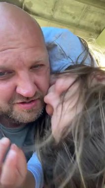 Bir adam ve genç bir kızın neşeli bir anı paylaştığı yakın plan bir fotoğraf. Kız erkeğin omzunda dinlenirken gülümsüyor, saçları rüzgarda uçuşuyor..