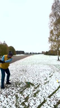 Kışın karla kaplı bir sahada, kalın giysiler içinde golf oynayan bir adam. Arka planda sonbahar ağaçları olan yerde kar parçaları var..