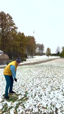 Kışın karla kaplı bir sahada, kalın giysiler içinde golf oynayan bir adam. Arka planda sonbahar ağaçları olan yerde kar parçaları var..