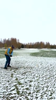 Kışın karla kaplı bir sahada, kalın giysiler içinde golf oynayan bir adam. Arka planda sonbahar ağaçları olan yerde kar parçaları var..