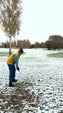 Kışın karla kaplı bir sahada, kalın giysiler içinde golf oynayan bir adam. Arka planda sonbahar ağaçları olan yerde kar parçaları var..