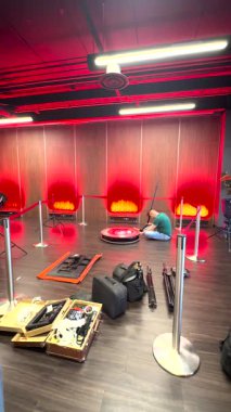 Riga, Letonya - 18 Ocak 2024 - İki kişi modern bir etkinlik alanında LED ışıkları olan bir 360 video kabini kuruyor. Ekipmanlar, kablolar ve çantalar etrafa saçılmış durumda..