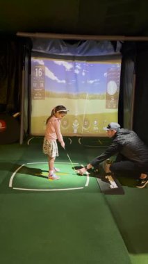 Valmiera, Letonya - 24 Kasım 2023 - Genç bir kız, yüksek teknoloji eğitim ortamında salıncakta salıncağında rehberlik alan, kapalı bir golf simülatöründe koçuyla golf oynuyor..