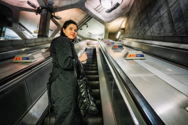 Londra, Birleşik Krallık - 23 Kasım 2022 - Yeraltı istasyonunda yürüyen merdiven üzerinde duran bir kadın, elinde bir su şişesi ve bir çanta, kameraya doğru bakıyor.
