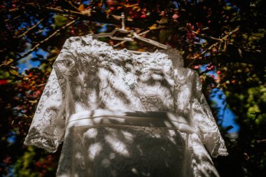 Ogre, Letonya - 11 Mayıs 2024 - Çiçeklerle çevrili beyaz dantelli bir gelinlik bir ağaç dalından sarkıyor, güneşin altında narin gölgeler bırakıyor.