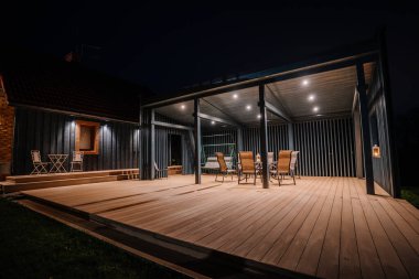 Geceleri modern bir ahşap teras, açık havada oturma, pergola ve sıcak ışıklandırma. Kapalı bir yemek alanı, salıncak ve dekoratif fenerler olan rahat bir ortam..
