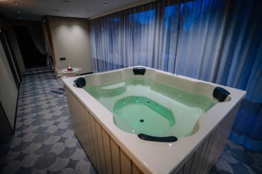 Suyla dolu lüks bir jakuzi, yumuşak ışıklandırmalı, zarif dekorlu, ve geniş perdeli, rahatlatıcı bir ortam yaratan rahat bir spa odası..