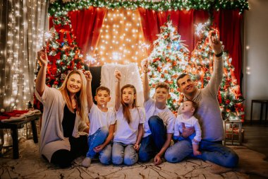 Gülümseyen aile maytap tutuyor, Noel ağaçları, ışıklar ve bayram neşesiyle süslenmiş bir odada oturuyor..