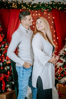 Gülümseyen çift birbirlerine sıcakkanlılıkla bakıyor. Parlayan ışıklar, süslenmiş ağaçlar ve paketlenmiş hediyelerle dolu bir Noel ortamında duruyorlar..