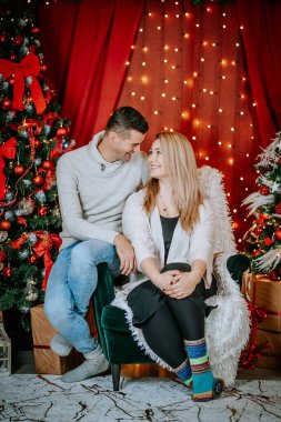 Sevgi dolu bir çift birbirine yakın oturuyor, samimi bir Noel ortamında gülümsüyor, neşeli ışıklar, ağaçlar ve etraflarına sarılmış hediyelerle..