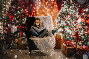 Çocuk rahat bir sandalyede kitap okuyor. Etrafında parlayan Noel ağaçları, kar örtüsü, tel lambalar ve paketlenmiş tatil hediyeleri var..