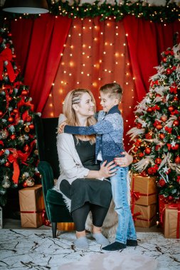 Sevgi dolu bir anne, ışıldayan ışıklar, süslenmiş ağaçlar ve arka planda paketlenmiş hediyelerle gülümseyen oğluna Noel ortamında sarılır..