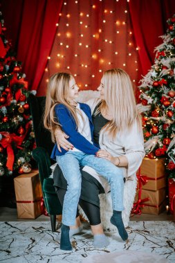 Anne ve kızı, süslenmiş ağaçlar ve ışıklarla süslenmiş bir Noel ortamında birlikte otururken birbirlerine içtenlikle gülümsüyorlar..