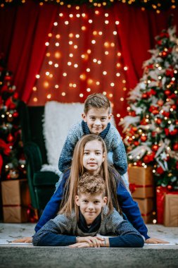 Üç gülümseyen çocuk neşeli bir poz içinde neşeli Noel ağaçlarının önünde parlayan ışıklarla ve kapalı mekanlarda hediyelerle..