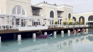 Belek, Türkiye - 25 Şubat 2025 - Bir alışveriş bölgesinde dekoratif şemsiyeler ve zarif mimari ile şık bir sahil restoranının altına kenetlenmiş gondol manzarası.