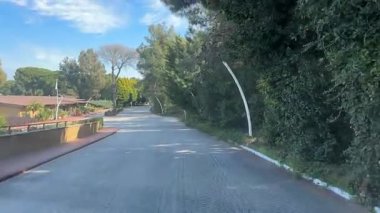 Belek, Türkiye - 25 Şubat 2025 - Bereketli, yeşil bahçeli, modern ışıklandırmalı ve solunda bir bina bulunan yeşil bir tatil köyünden geçen huzurlu bir asfalt yol.