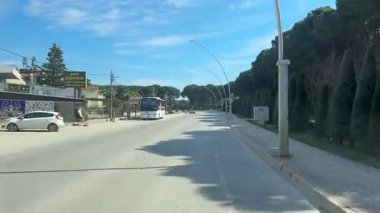 Belek, Türkiye - 25 Şubat 2025 - Bir dağıtım kamyonu, park etmiş arabalar, dükkanlar ve uzakta bir otobüsle gün ışığında bir aracın ön camından yakalanan güneşli yol kenarı sahnesi.