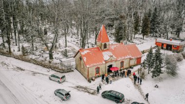 Valmiera, Letonya - 6 Ocak 2022 - Kırmızı çatılı küçük bir kilisenin havadan görünüşü ve insanlar karlı bir kış günü, bir orman ve mezarlıkla çevrili olarak dışarıda toplandılar..