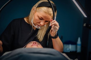 Güzellik uzmanı, profesyonel güzellik salonu ortamında rahat bir müşteriye kirpik uzatma işlemi yaparken büyüteç ayarlıyor..