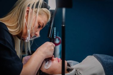 Bir güzellik uzmanının yakınlaştırma ışığı altında cımbızla kirpik eklentileri uygularken müşteri spa ortamında göz pedleri ile rahatlarken.