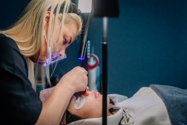 Güzellik uzmanı, bir kuafördeki müşteriye kirpik eklentisi uygularken ultraviyole ışık kullanıyor, işlem sırasında hassasiyet ve hijyene odaklanıyor..