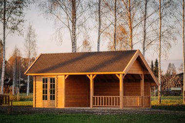Ahşap bahçe kulübesi, çatısı ve verandası ile kaplı, sakin bir sonbahar manzarası, huş ağaçları ve yumuşak akşam güneş ışığı ile kurulmuş.