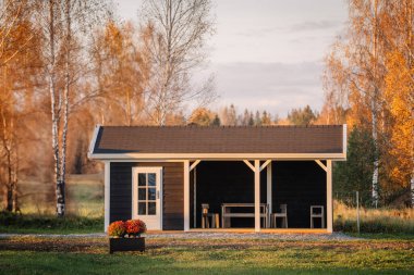Beyaz bukleli küçük, modern, siyah ahşap bir kulübe huzurlu bir sonbahar manzarasında duruyor, verandasında mobilyalar ve önünde çiçekler olan bir saksı..