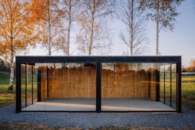 Modern cam bahçe binası, siyah bir çerçeve ve ahşap arka duvarı ile, sonbaharda huş ağaçları arasında inşa edilir sıcak doğal aydınlatma ve çakıl temeli ile.