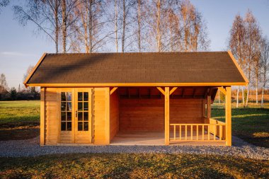 Valmiera, Letonya - 2 Kasım 2023 - Kiremit çatılı ve küçük verandalı ahşap bahçe evi, sıcak güneş ışığı ile aydınlatılmış, çıplak huş ağaçları ile çevrili huzurlu bir kırsal alanda.