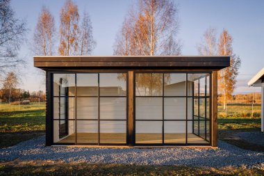 Siyah metal çerçeveli modern minimalist cam kulübe, huş ağaçlarıyla çevrili sonbahar manzarasını ve altın saat boyunca açık araziyi yansıtıyor..