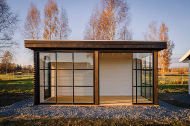 Valmiera, Letonya - 2 Kasım 2023 - Siyah çerçeveli ve sürgülü kapılı modern cam bahçe evinin ön görüntüsü, sonbahar manzarasında ağaçlar ve yumuşak altın güneş ışığı.