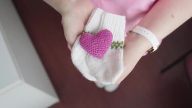 Valmiera, Letonya - 24 Mart 2025 - Küçük mor bir tığ işi kalp ile beyaz örgü bebek çorapları tutan bir kişinin yakın çekimi, ilgi, sevgi ve yeni doğan.