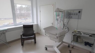 Valmiera, Letonya - 24 Mart 2025 - Yeni doğan bakım ekipmanları, dijital bebek terazisi, siyah sandalye ve park alanına bakan bir pencereyle dolu parlak bir hastane odası.