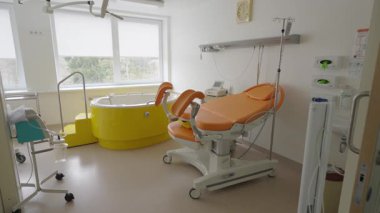Valmiera, Letonya - 24 Mart 2025 - Sarı doğum küveti, turuncu doğum yatağı, tıbbi cihazları ve doğal ışık için büyük pencereleri olan parlak ve modern doğum odası.