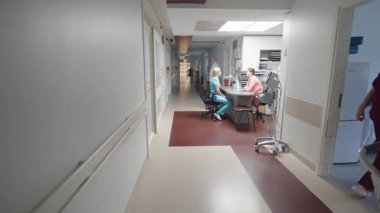 Valmiera, Letonya - 24 Mart 2025 - Tıp personeli, bilgisayar, dosya ve tıbbi ekipmanlarla dolu bir hemşire istasyonunun yanındaki parlak ışıklı hastane koridorunda yürür ve yürür..