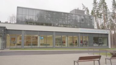 Valmiera, Letonya - 24 Mart 2025 - Geniş cam pencereli ve görünür iç mekanlı modern rehabilitasyon merkezi, çevresi çam ağaçları ve banklarla çevrili barışçıl bir açık alan.