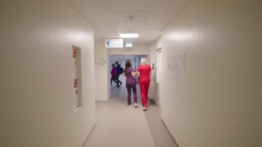 Valmiera, Letonya - 24 Mart 2025 - Renkli önlüklü iki sağlık çalışanı temiz bir hastane koridorundan daha aydınlık bir ortak alana doğru yürüyorlar..