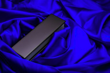 Zarif kıvrımlı, koyu mavi saten kumaşa yerleştirilmiş parlak siyah dikdörtgen kutu, kaliteli ürün gösterimi, markalaşma veya ambalaj tasarımı için ideal..