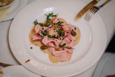 Riga, Letonya - 27 Mart 2025 - Kremalı sos, kapari üzümü ve taze otlarla servis edilen zarif et dilimleri gurme yemek ortamında beyaz porselenler üzerine sunulmuştur..