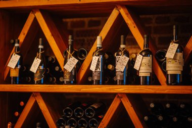 Riga, Letonya - 27 Mart 2025 - Kırsal bir şarap mahzenindeki ahşap raflarda el yazısıyla yazılmış çeşitli şarap şişeleri sıcak ve zarif bir vitrin oluşturuyor..