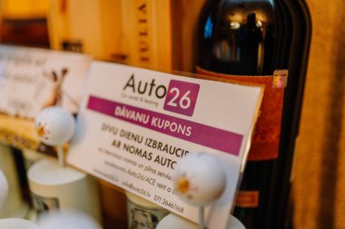Riga, Letonya - 27 Mart 2025 - Golf kupaları ve şarap şişeleri arasına yerleştirilmiş bir Auto26 hediye çeki, kapalı bir mekanda kiralık araba deneyimini destekliyor.