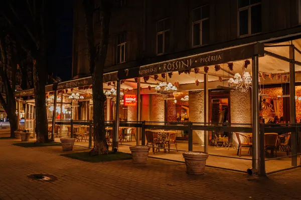 Riga, Letonya - 27 Mart 2025 - Rossini restoranı ve pizzacının gece manzarası sıcak ışıklar, cam duvarlar ve zarif bir ambiyansı olan sakin bir sokakta açık hava koltukları.