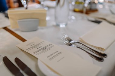 Riga, Letonya - 27 Mart 2025 - İtalyan menüsü, gümüş takımı, peçete ve su şişesi olan bir restoranın güzel bir yemek ortamında beyaz bir masa örtüsü üzerine kurulumu.