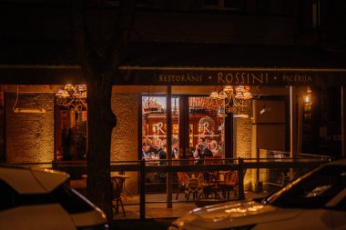 Riga, Letonya - 27 Mart 2025 - Rossini restoranı geceleri sıcak bir şekilde aydınlatılıyor, misafirler içeride yemek yiyor, büyük pencerelerden görülebiliyor, rahat ortam ve zarif ışıklandırmayla çevrili..