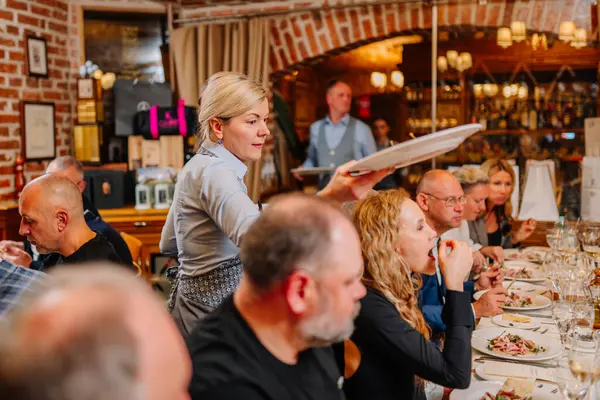 Riga, Letonya - 27 Mart 2025 Garson, sıcak bir restoranda bir grup restorana yemek servisi yapıyor. Misafirler sıcak ve samimi bir yemek atmosferinde resmi yemeğin tadını çıkarıyorlar..