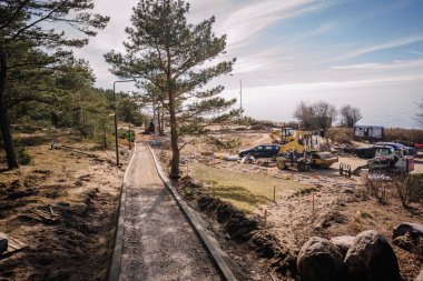 Salacgiva, Letonya - 27 Mart 2025 - Güneşli bir günde arka planda makine, çakıl yolu, çam ağacı ve okyanus bulunan deniz kenarı şantiyesi.