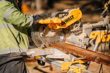 Salacgiva, Letonya - 27 Mart 2025 - DeWalt miter testeresiyle tahta kesen işçiler, gün ışığında bir inşaat alanında talaş üretiyorlar..