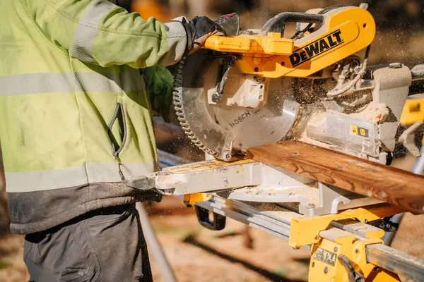 Salacgiva, Letonya - 27 Mart 2025 - DeWalt miter testeresiyle tahta kesen işçiler, gün ışığında bir inşaat alanında talaş üretiyorlar..