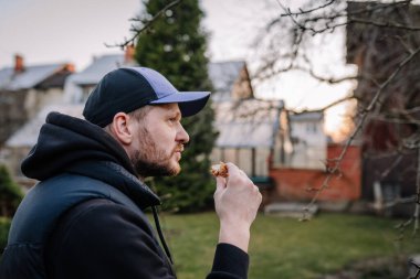Valmiera, Letonya - 29 Mart 2025 - Soğuk hava mevsiminde arka planda yapraksız ağaç dalları ve evleri olan bir bahçede günlük kıyafet ve kep giyen bir adam açık havada yemek yiyor.