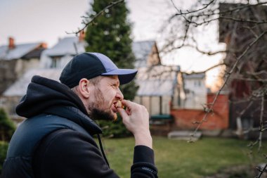 Kapüşonlu ve şapkalı bir adam serin bir günde arka planda ağaçlar ve binalar olan bir konut bahçesinde dikilirken bir lokma yemek yiyor..